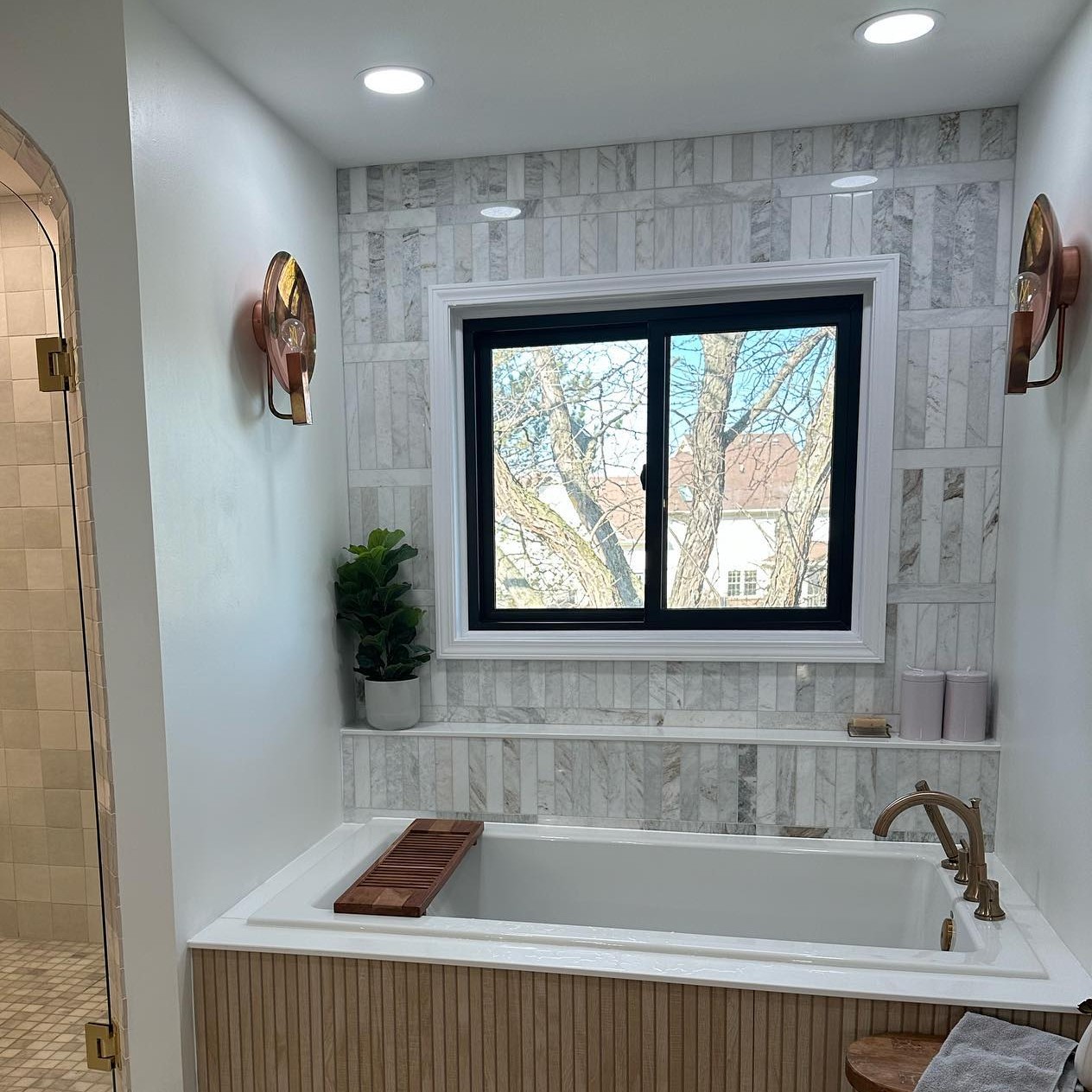 A homeowner sent us this photo of their bathroom remodel. It has luxe white marble and brass fixtures and a black gliding or sliding window above the bathtub.
