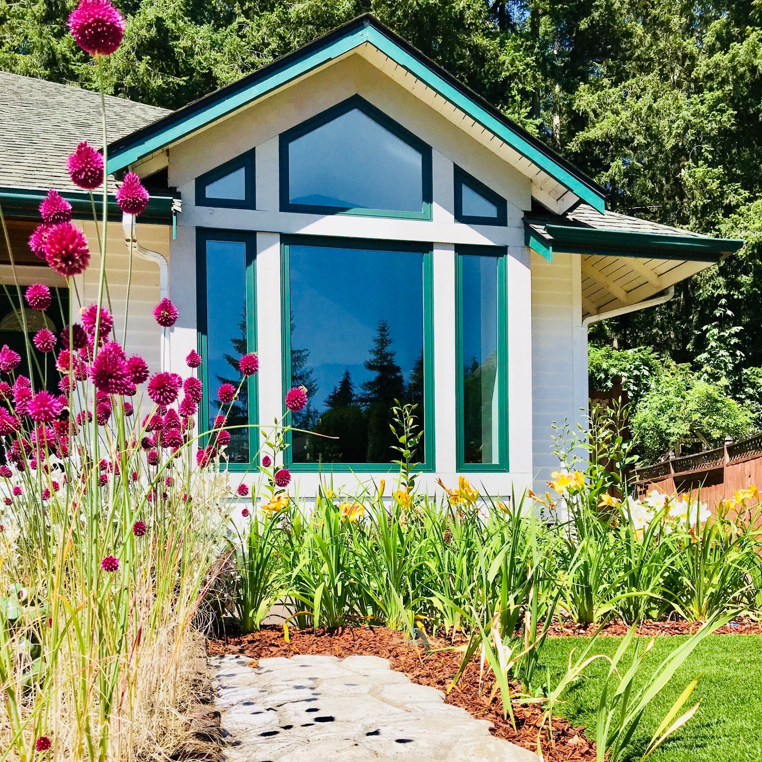 Two small trapezoid windows flank a peak pentagon window. These are set atop a bank of picture windows. The windows are forest green frames. There are pink flowers in the foreground providing contrast.