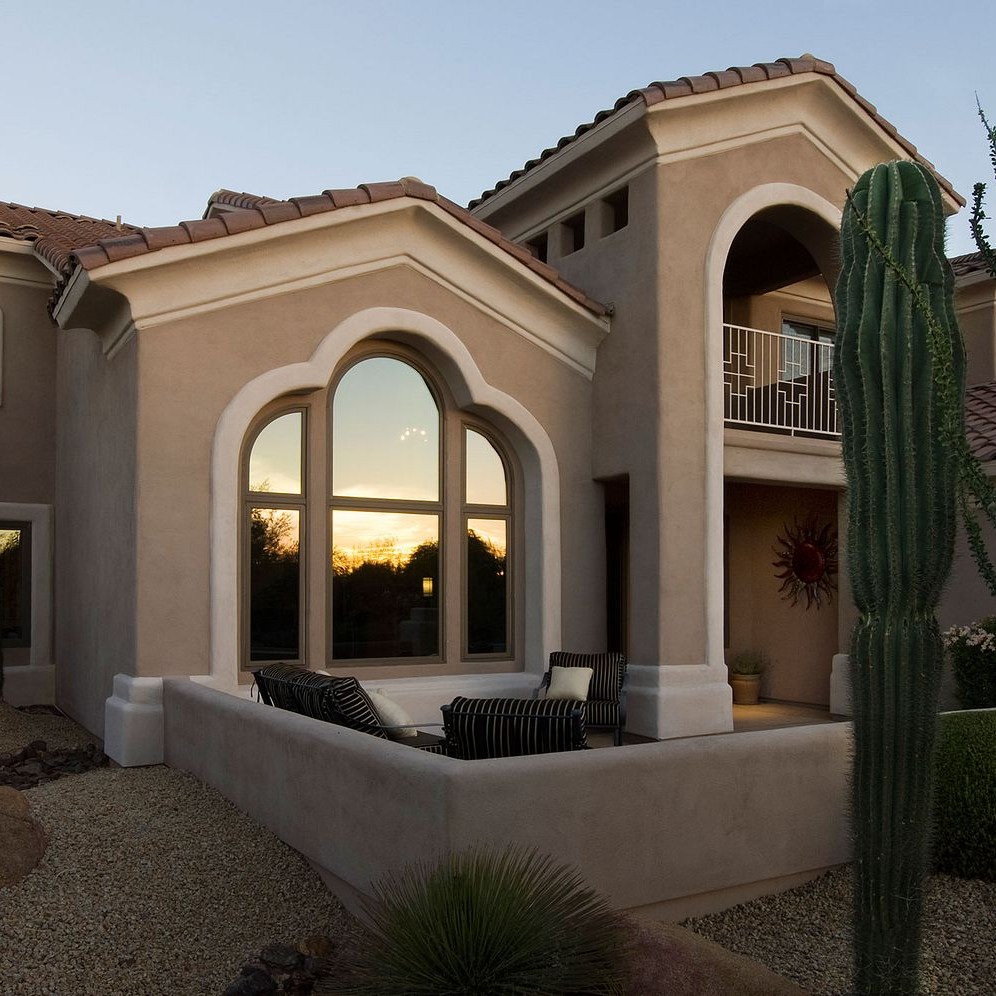 An adobe style home in the southwestern desert. The sunset is being reflected in the windows as twilight approaches. The home has picture windows topped with quarter circle and springline windows.